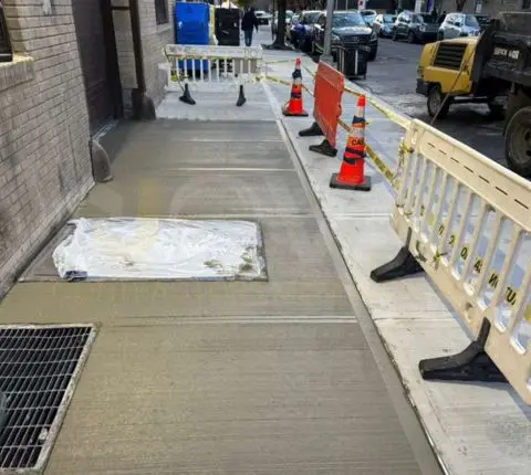 Newly poured sidewalk beside a building entrance, protected by safety barriers and cones