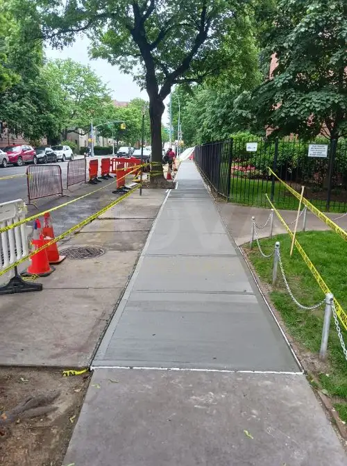 Newly replaced concrete sidewalk along a tree-lined street, with safety barriers around the work zone
